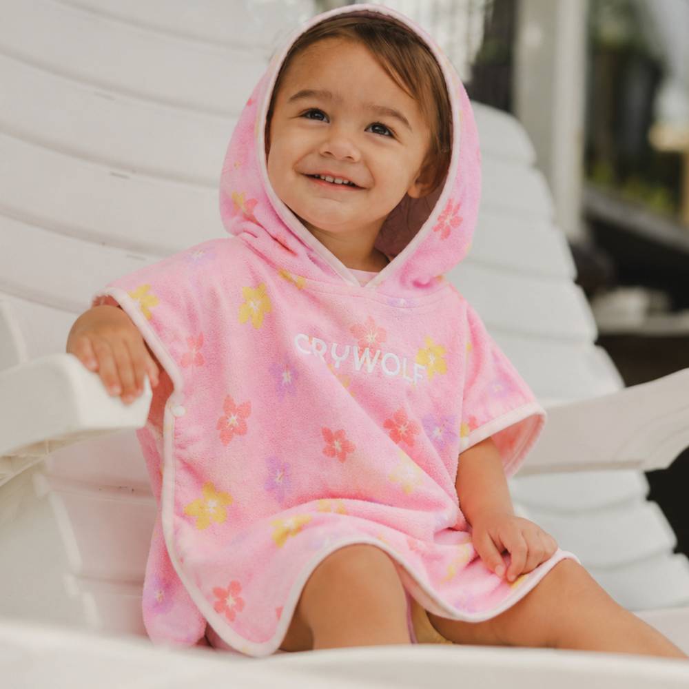 A smiling young child wearing a pink hooded baby towel with colorful floral print and the brand name 'CRY WOLF' on the front, seated on a white outdoor chair.