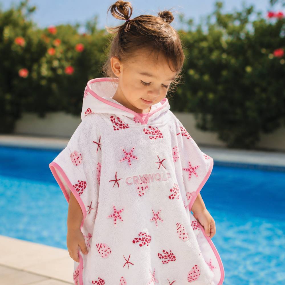 A toddler wearing a white hooded towel with pink trim and red starfish and shell prints, standing by a swimming pool outdoors. The brand name CRYWOLFis printed on the front.