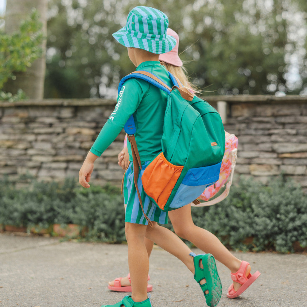 A child walking outdoors wearing a green and blue striped sun hat, a green long-sleeve rash guard with blue trim, matching striped swim shorts, a green backpack with orange and blue side pockets, and green clogs.