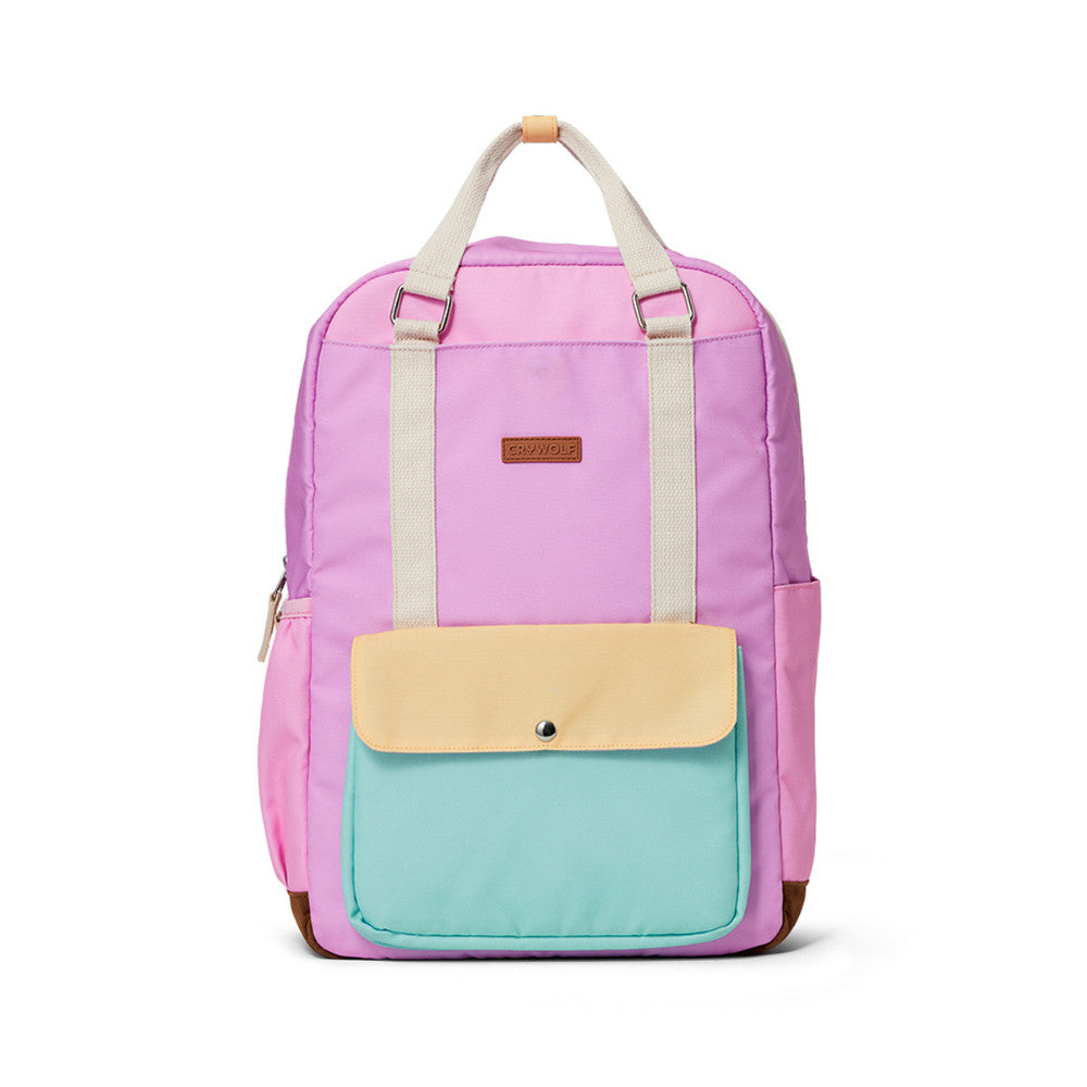 A pastel color-blocked backpack with a purple upper body, a mint green front pocket, and a pale yellow flap, featuring cream-colored top handles and a small brown brand patch.