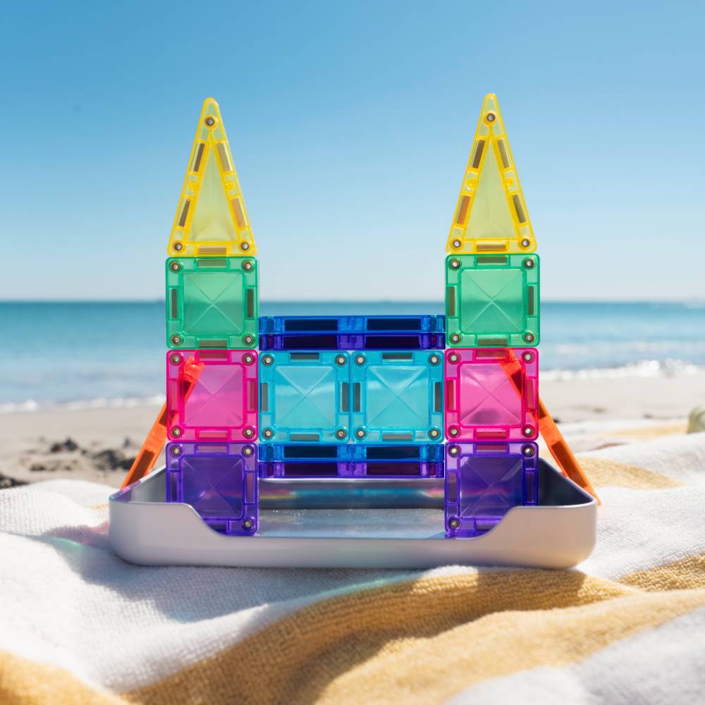 Colorful magnetic building blocks arranged in a castle-like structure on a beach blanket with the ocean and blue sky in the background.