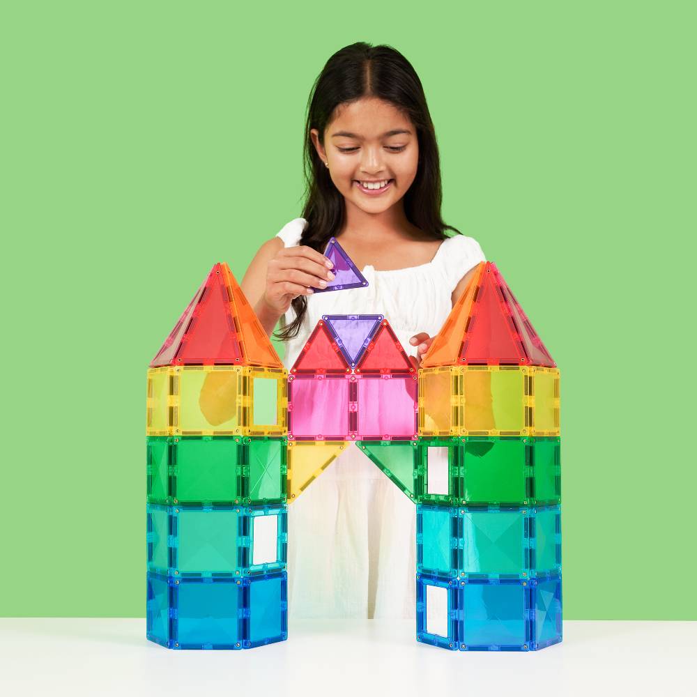 A young girl with long dark hair, wearing a white top, smiles while building a colorful translucent block castle with a purple triangular piece, set against a solid light green background.