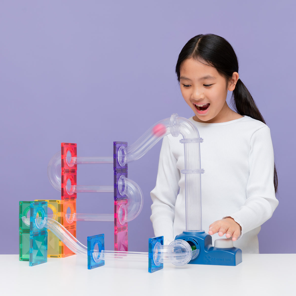 A young girl with dark hair in a ponytail, wearing a white long-sleeve shirt, is smiling and playing with a colorful translucent acrylic marble run toy set on a white table against a solid light purple background.