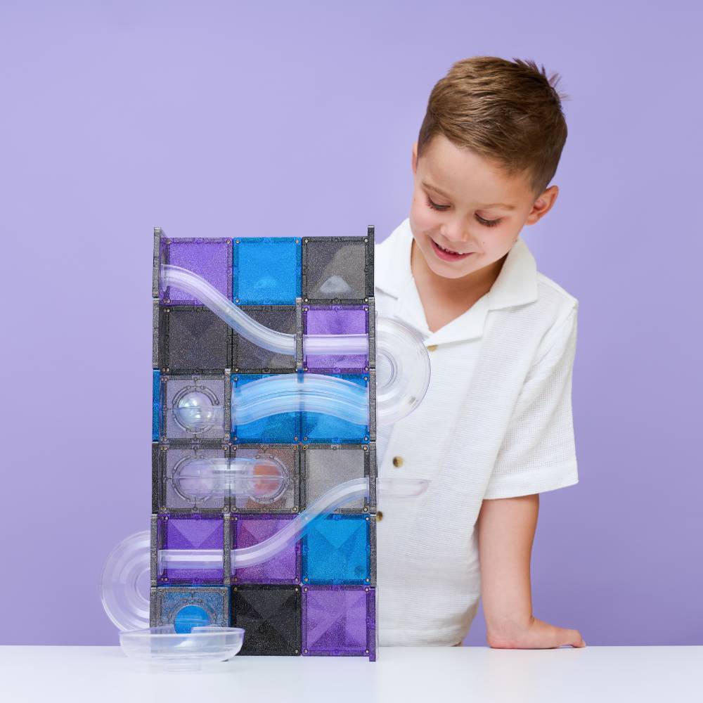 A young boy in a white polo shirt smiles while looking at a magnetic ball run toy built from purple, blue, and black translucent tiles on a white table against a solid lavender background.