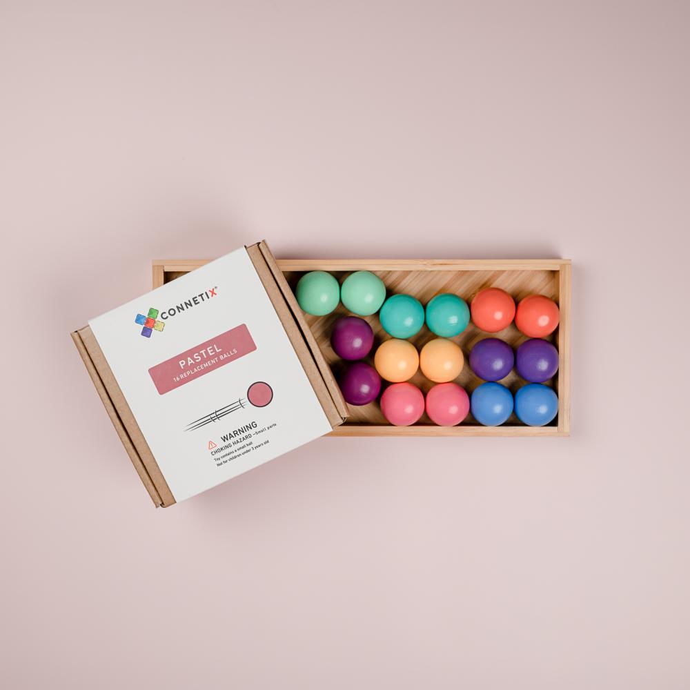 A wooden box of pastel balls by Connetix, with the lid showing the product packaging and the open tray filled with colorful pastel balls on a soft pink background.