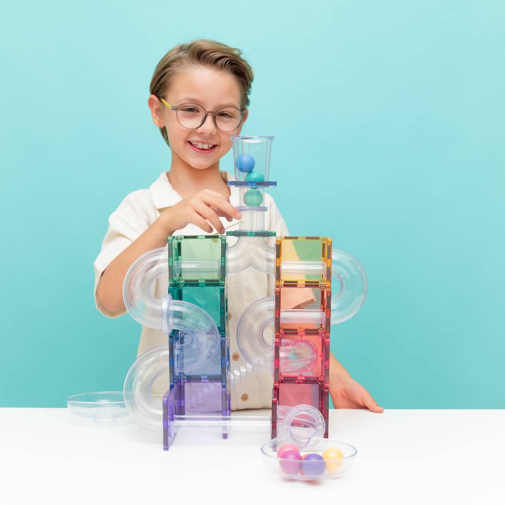 A young boy with glasses is smiling and playing with a transparent acrylic ball run toy set on a white table against a light blue background. The colorful magnetic tiles and balls are arranged in a complex track, with bowls of small marbles on the table.