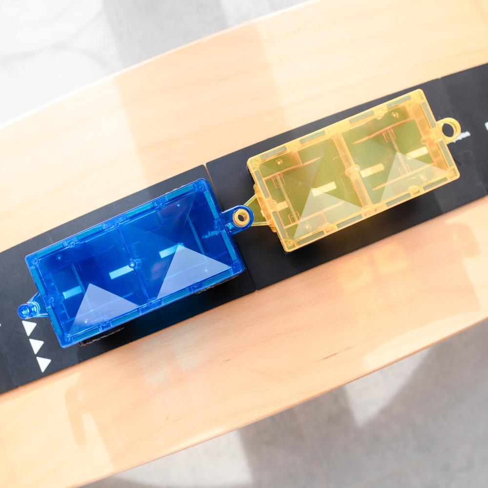 Two toy trucks, one blue and one yellow, on a black surface with a wooden background.