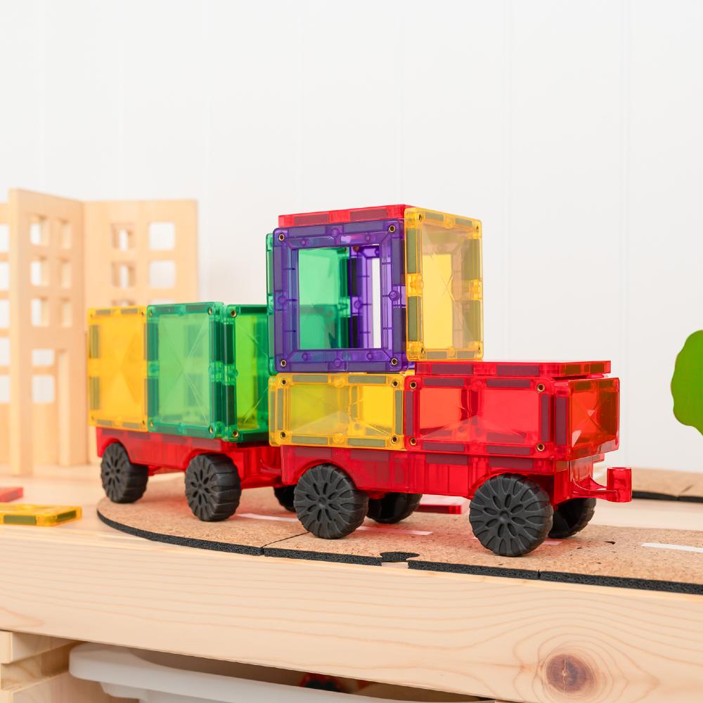 A colorful magnetic building toy train set on wooden tracks, featuring a red train car carrying stacked translucent cargo containers in yellow, green, purple, and red, with black wheels.