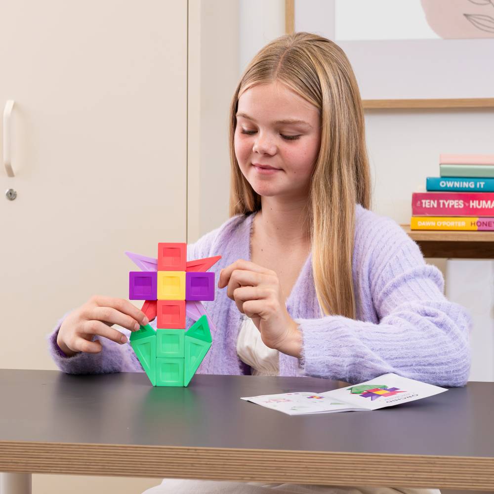 A young girl with blonde hair sits at a wooden desk, assembling a colorful plastic building toy that resembles a flower. She is wearing a light purple knit cardigan.