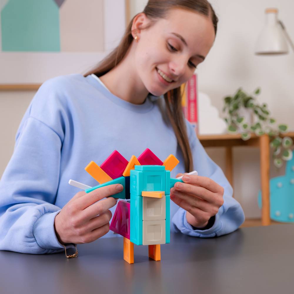A young lady in a light blue sweatshirt smiles while assembling a colorful geometric toy structure with teal, orange, pink, and beige magnetic tiles on a dark tabletop in a bright indoor setting.