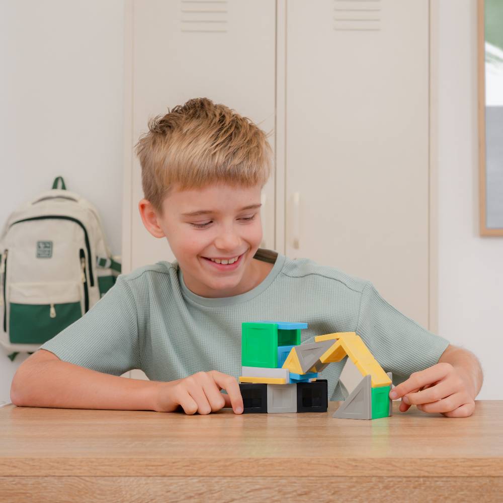 A young boy with blonde hair smiling while playing with colorful geometric magnetic tiles on a wooden table in a classroom setting.