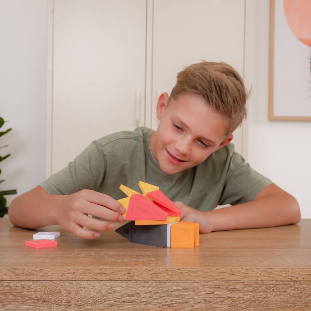 A young boy with blonde hair wearing an olive green t-shirt is sitting at a wooden table, smiling as he plays with colorful geometric magnetic tiles.