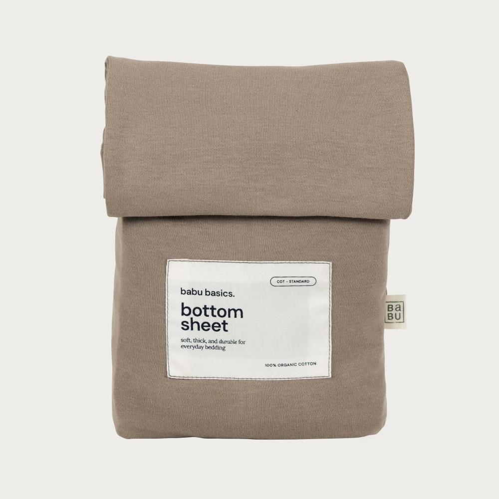 A folded bottom sheet in a warm taupe-beige color, featuring a white label that reads 'babu basics. bottom sheet' and a small side tag, set against a solid off-white background.