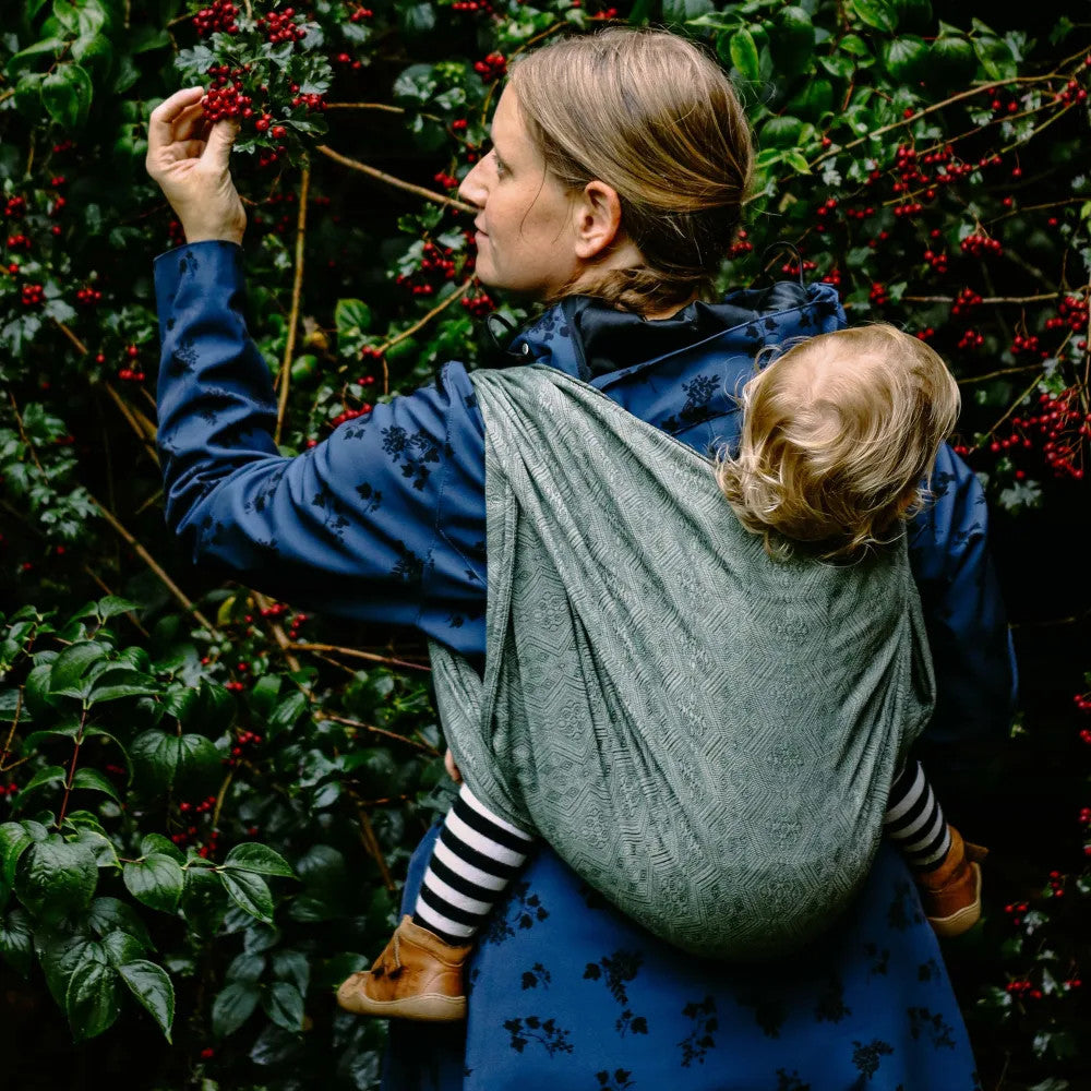 Woman wearing a Baby Wrap Sling Linen - Whispering Woods on her back with toddler inside. 