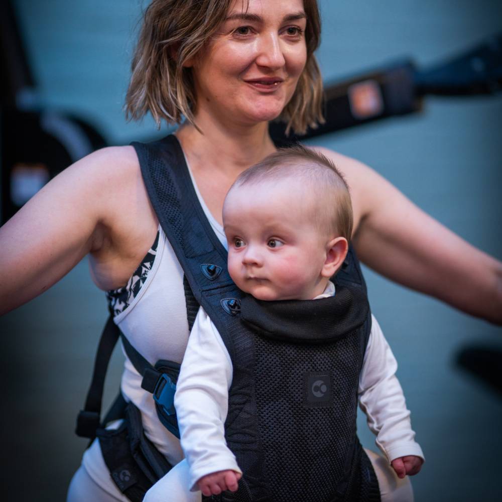 Woman carrying a baby in a black baby carrier against a blurred background