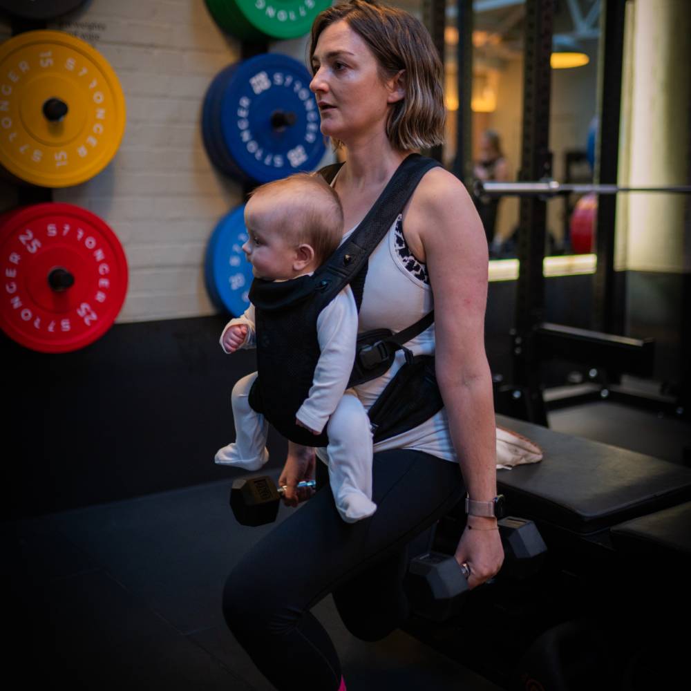 Woman holding a baby in a Carifit baby carrier while exercising in a gym.
