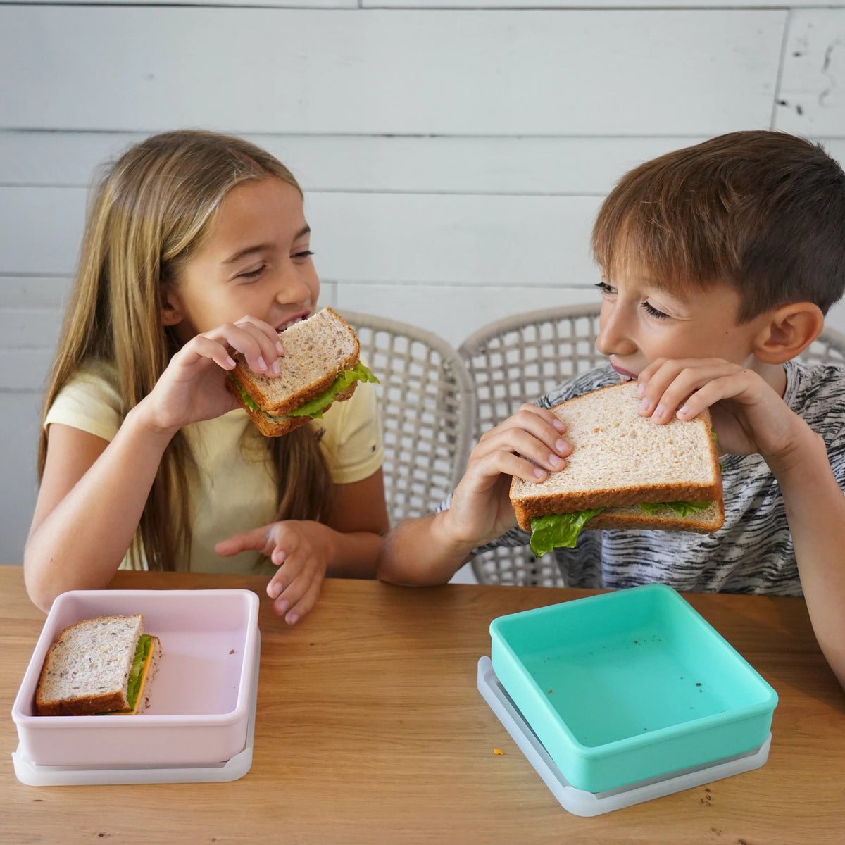 Two children eating lunch at a wooden table, each holding a sandwich half with a reusable container lid nearby.