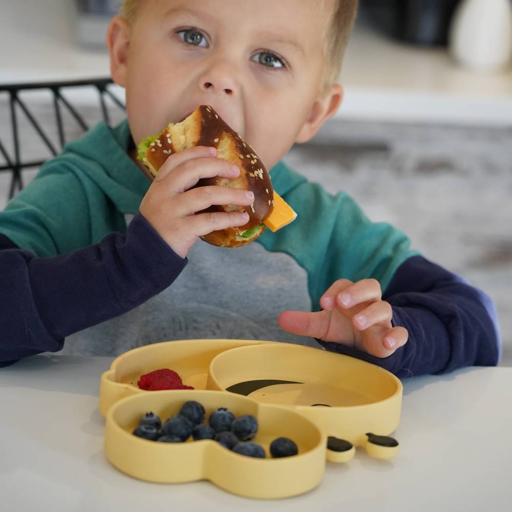 Child eating a sandwich with a yellow divided bee shaped plate containing fruits on a table.