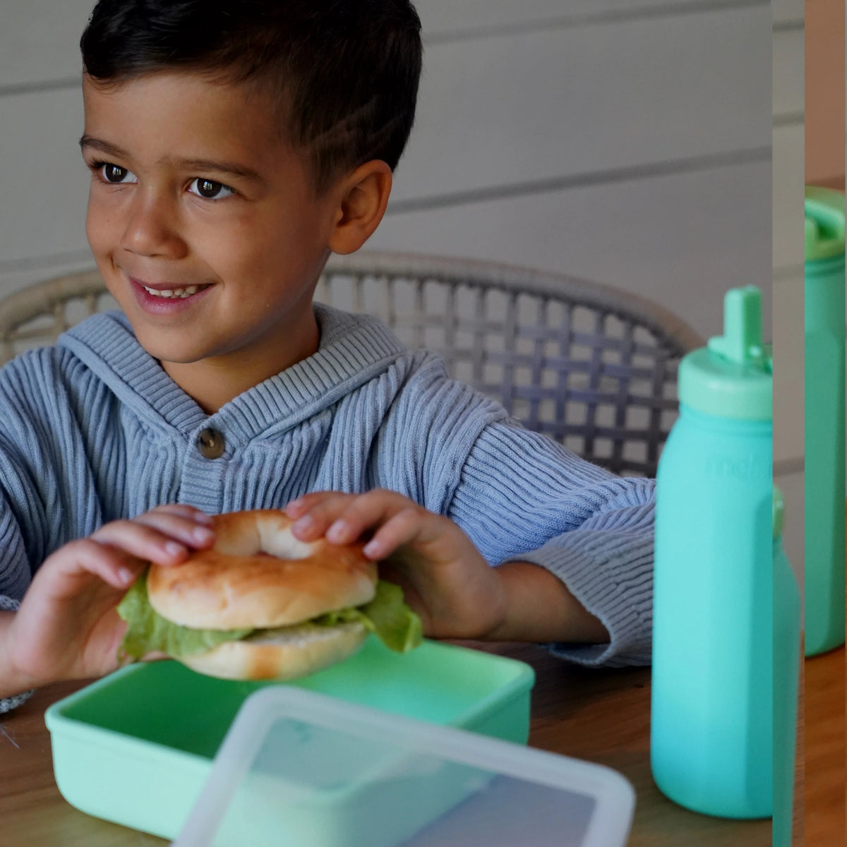 A young boy in a blue striped shirt smiles while holding a bagel with lettuce. In front of him on a wooden table is a mint green lunch container with a clear lid, alongside a matching mint green water bottle.