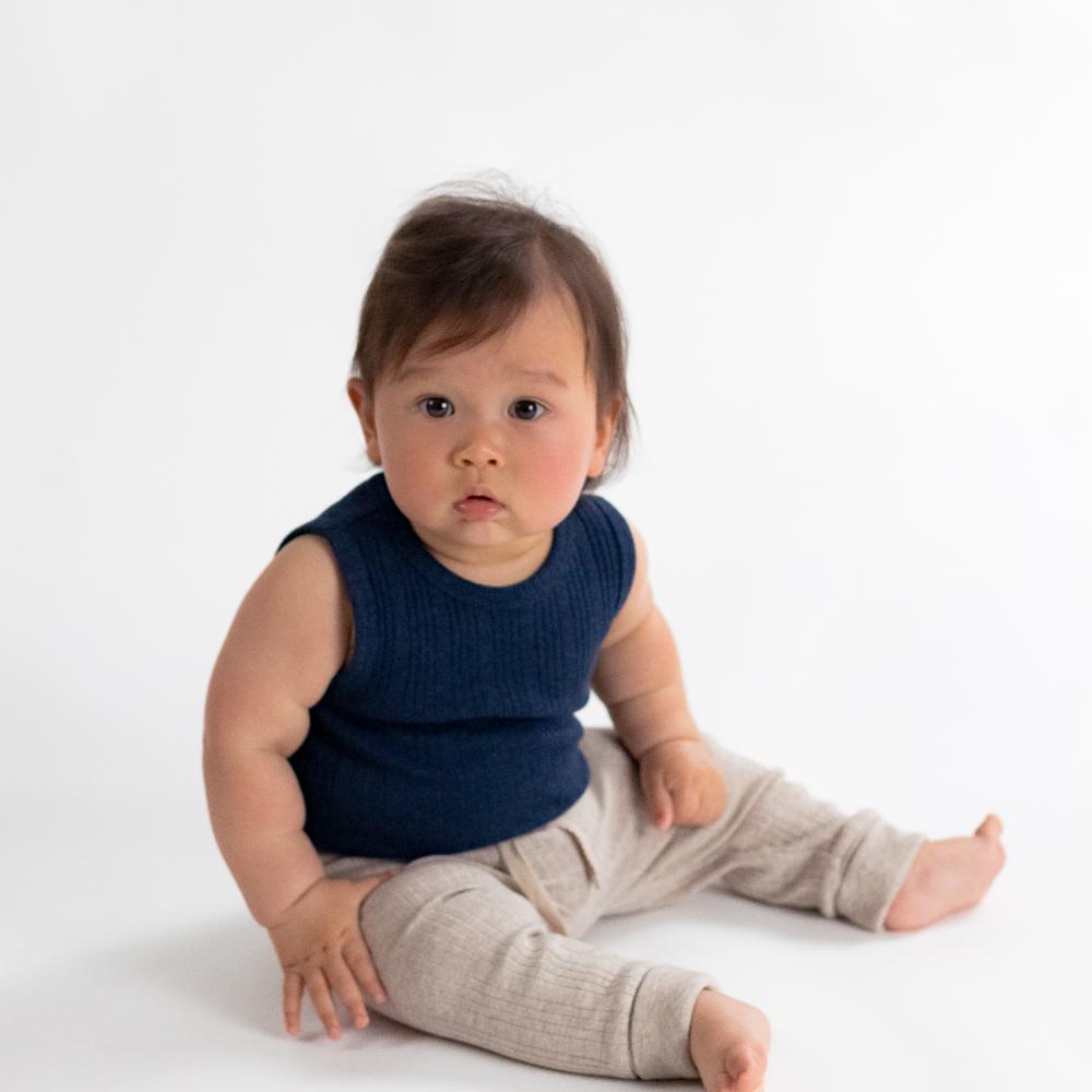 Baby sitting on a white background wearing a blue singletsuit and beige pants.