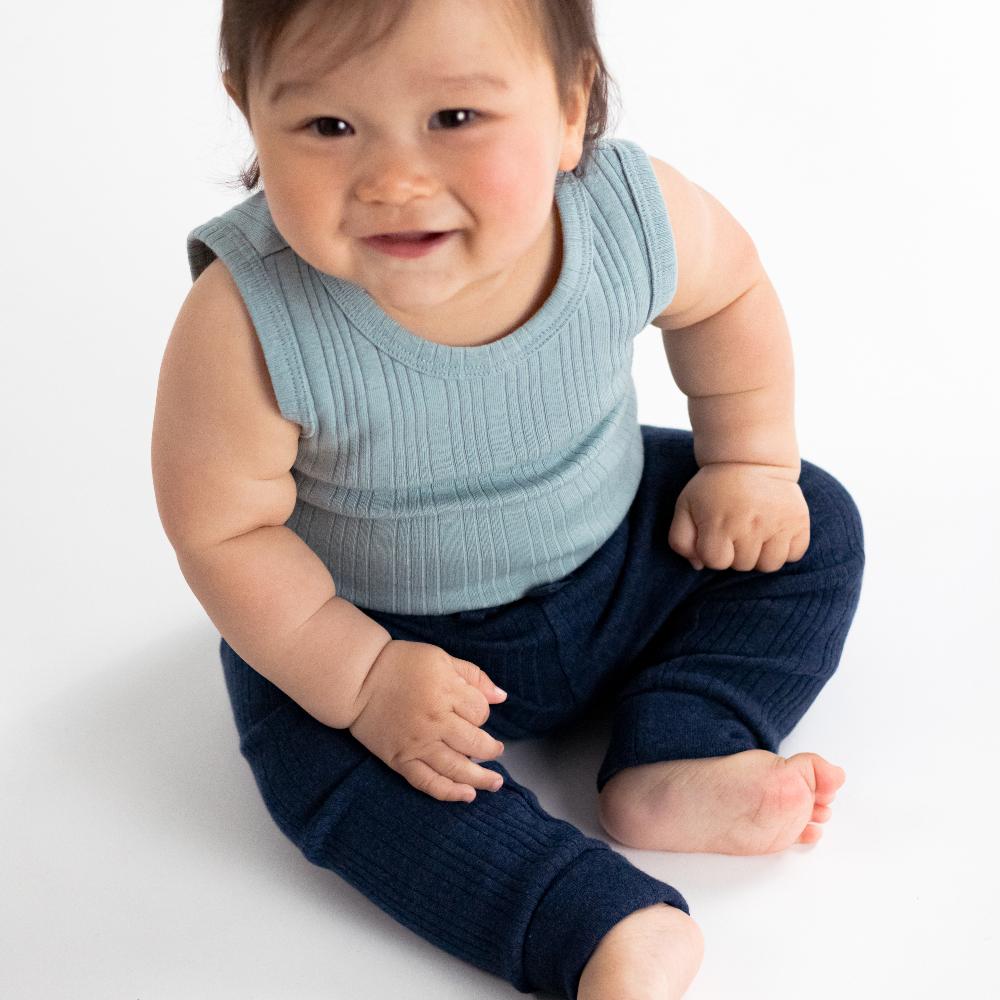 Baby wearing a light blue sleeveless top and dark blue merino pants on a white background