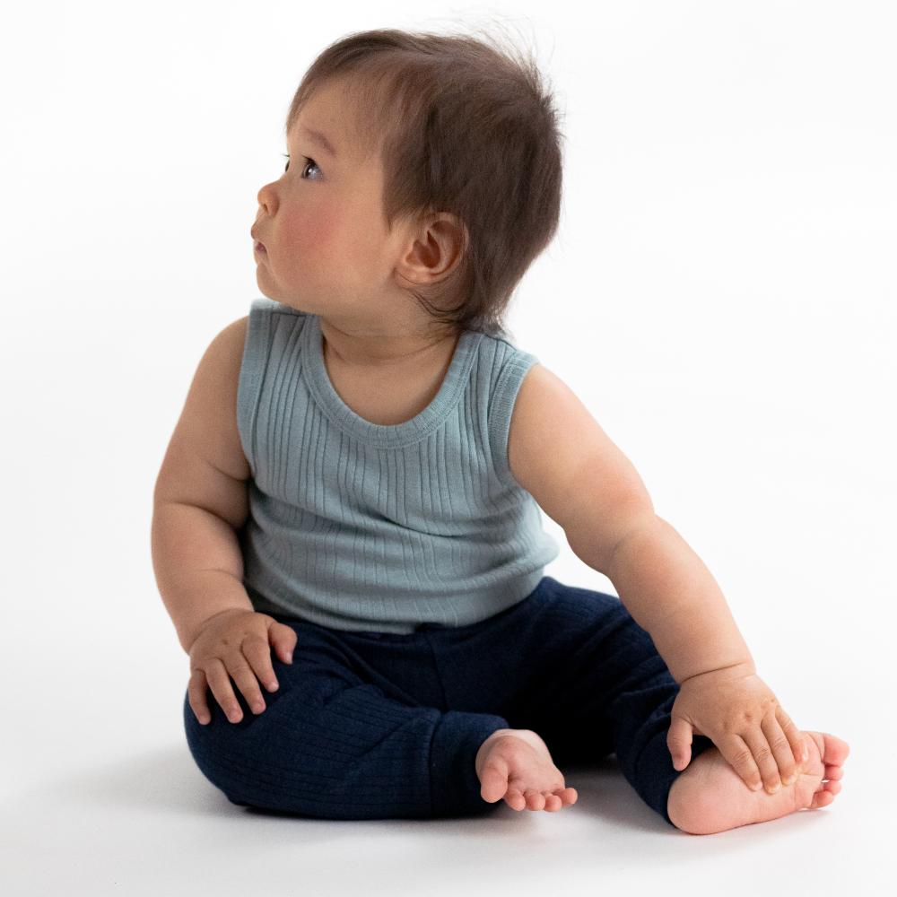 Baby sitting on a white surface wearing a light blue sleeveless shirt and dark blue woolbabe baby pants.