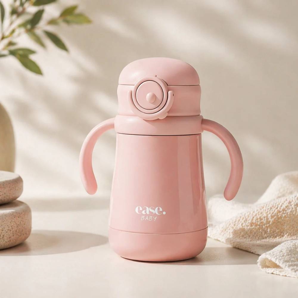 A light pink baby water bottle with integrated side handles and a flip-top lid, branded 'ease. baby', set on a warm beige surface with soft shadows and decorative props.