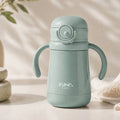 A light sage green baby water bottle with integrated side handles and a flip-top lid, featuring the 'ease. baby' logo on the front, set against a warm, softly lit background with decorative props.