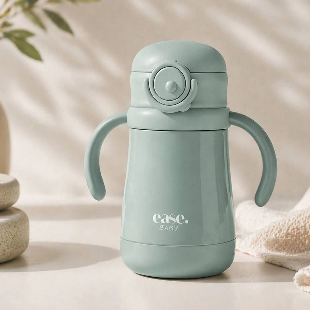 A light sage green baby water bottle with integrated side handles and a flip-top lid, featuring the 'ease. baby' logo on the front, set against a warm, softly lit background with decorative props.