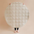 A person holding up a very large round quilted mat or pad in a studio setting. The product is light cream or off-white with a stitched square grid pattern.