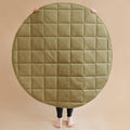 A large round quilted floor mat or play mat in an olive green color, held up by a person whose legs and bare feet are visible against a solid beige background.
