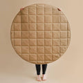 A large round quilted tan floor mat or rug held up by a person whose hands and bare feet are visible against a plain beige background.