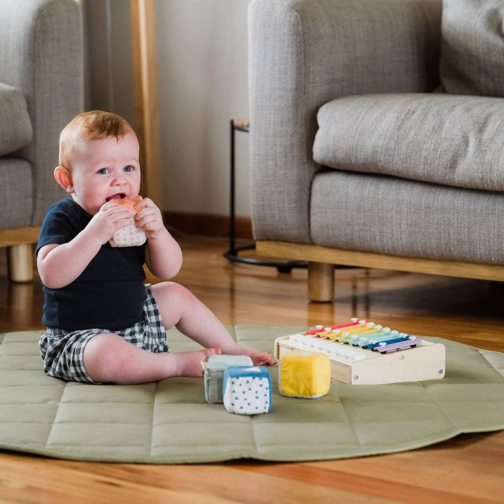 A baby sits on a round quilted play mat on a wooden floor in a living room, holding a toy cup. The play mat is accompanied by wooden blocks and a xylophone toy.