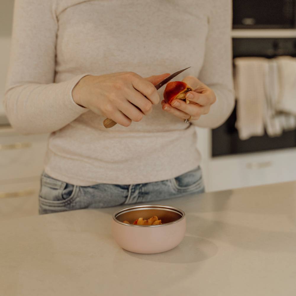 A person in a light beige sweater and blue jeans stands at a kitchen counter, peeling a peach with a small knife while a blush pink stainless steel bowl sits in front of them.