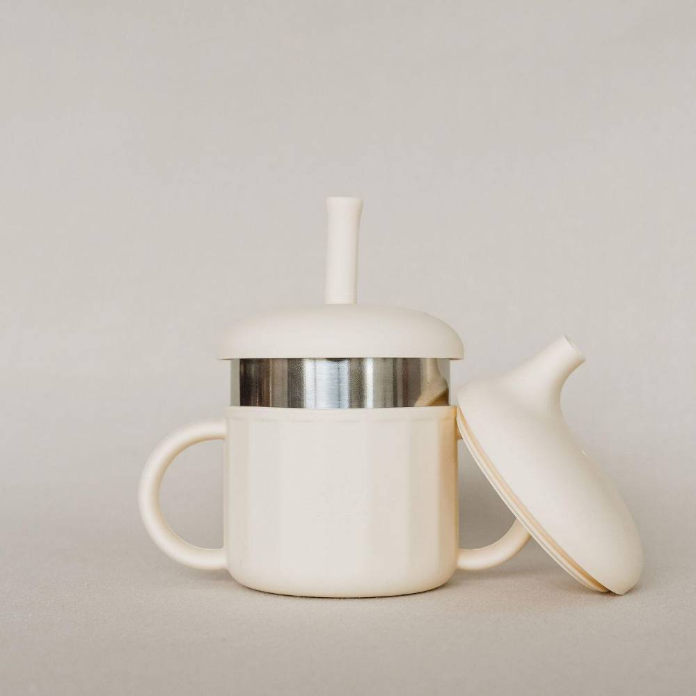 An off-white silicone and stainless steel baby sippy cup with a matching lid resting beside it, featuring a stainless steel band around the middle and a small handle on the left side, set against a neutral light gray background.