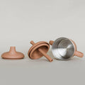 A terracotta-colored silicone baby bottle lid and straw set displayed next to an open stainless steel bottle with a matching terracotta handle, on a neutral light gray background.