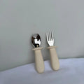 A baby spoon and fork set with matte beige handles and polished silver metal heads, standing upright on a white textured surface against a light gray wall.