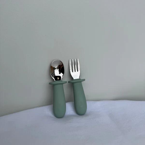 A baby utensil set featuring a spoon and a fork with matte sage green silicone handles and polished stainless steel heads, standing upright against a neutral gray background.