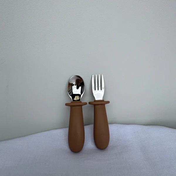 A baby feeding utensil set consisting of a spoon and a fork with matte brown silicone handles and silver metal heads, standing upright on a light gray fabric surface against a matching gray wall.