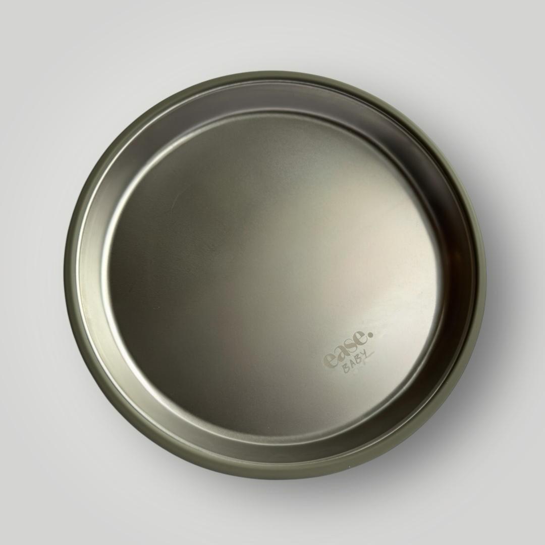 A round stainless steel plate viewed from directly above on a light gray background, with the brand name 'ease.baby' subtly embossed near the bottom right.