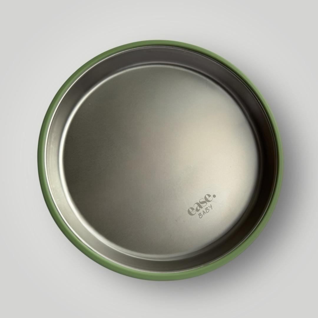 Top-down view of a round stainless steel plate with a green rim, showing the embossed 'ease.baby' logo on a light gray background.