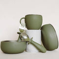 An olive green silicone baby feeding set by ease.baby, including a silicone cup, bowl, spoon, and fork, arranged on a white cylindrical pedestal against a light beige background.