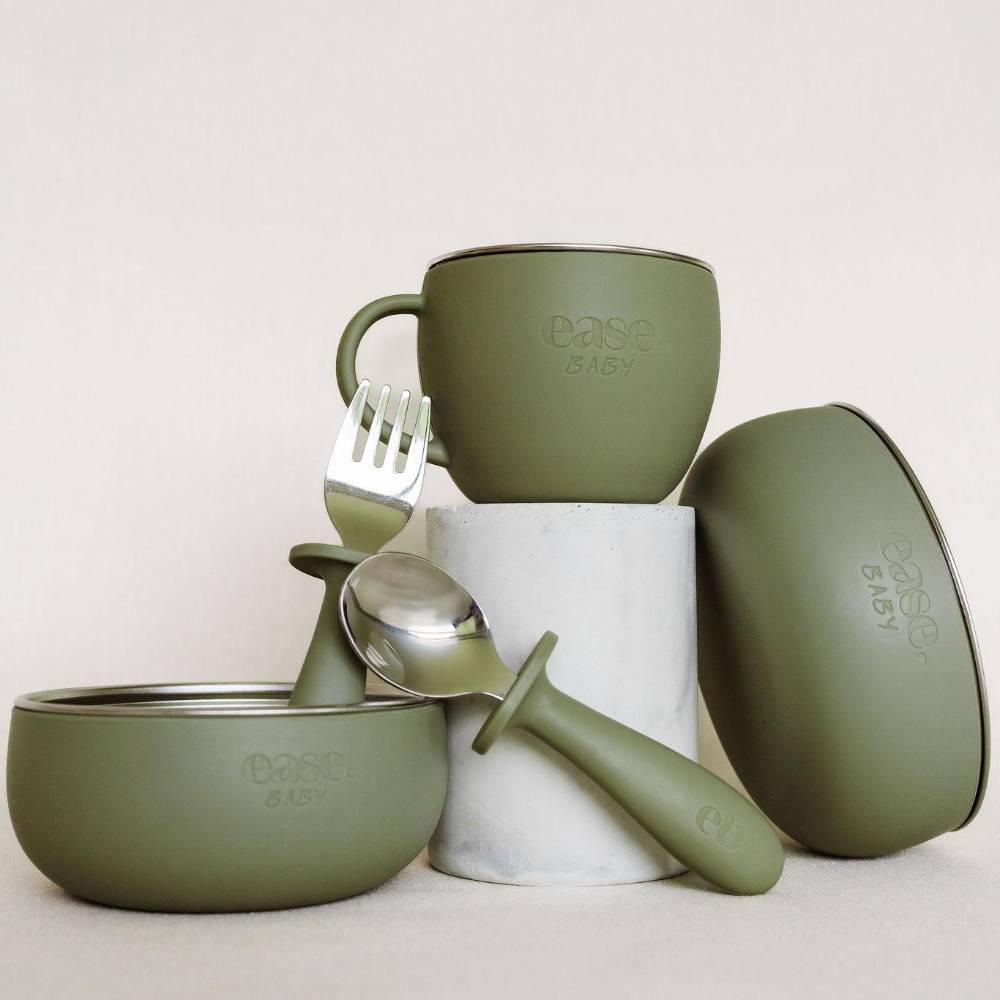 An olive green silicone baby feeding set by ease.baby, including a silicone cup, bowl, spoon, and fork, arranged on a white cylindrical pedestal against a light beige background.