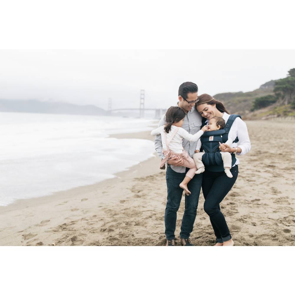 A family of four on a sandy beach with the Golden Gate Bridge visible in the misty background. The mother is wearing a dark navy baby carrier with a baby inside, while the father holds a toddler.