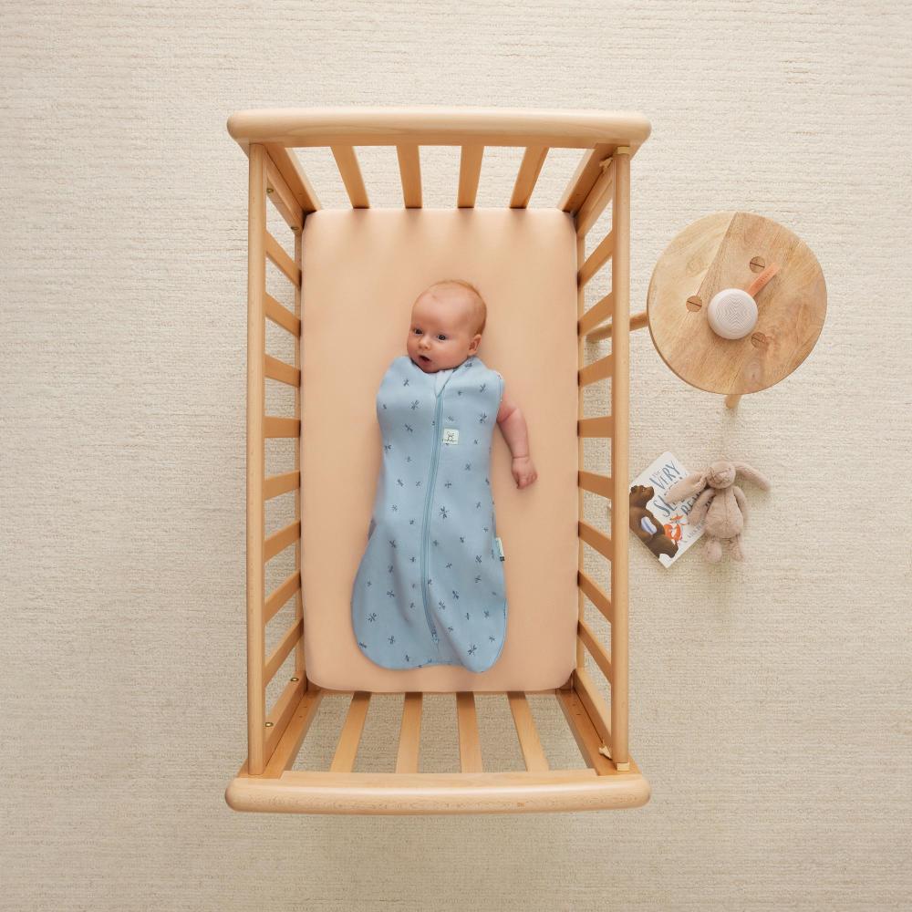 Baby in a blue swaddle with one arm out lying in a wooden crib with a beige mattress.