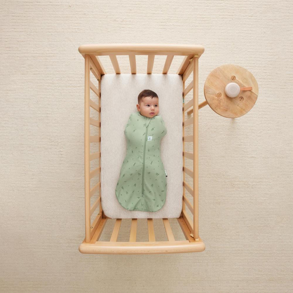 Baby in a green outfit lying in a wooden crib with a beige wall background