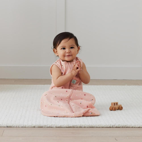 Baby in a pink sleep sack sitting on a white mat with a wooden toy car.