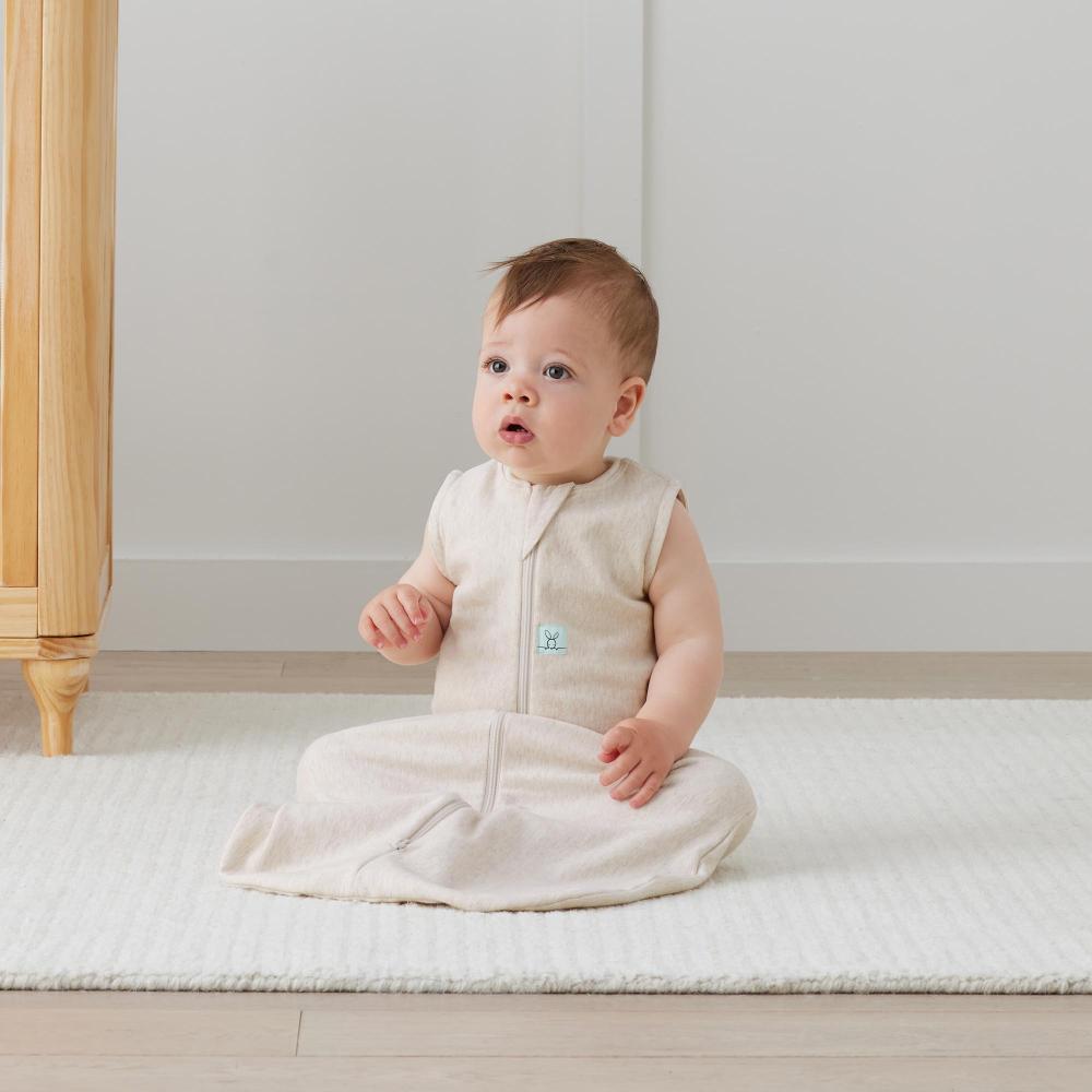 Baby sitting on a white mattress wearing a beige sleeveless outfit with a small logo.