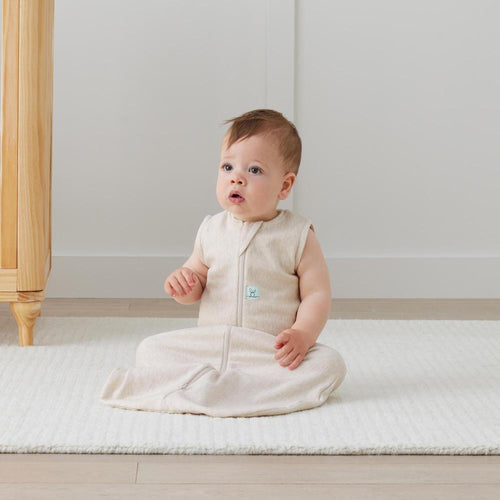 Baby sitting on a white mattress wearing a beige sleeveless sleep sack with a small logo.