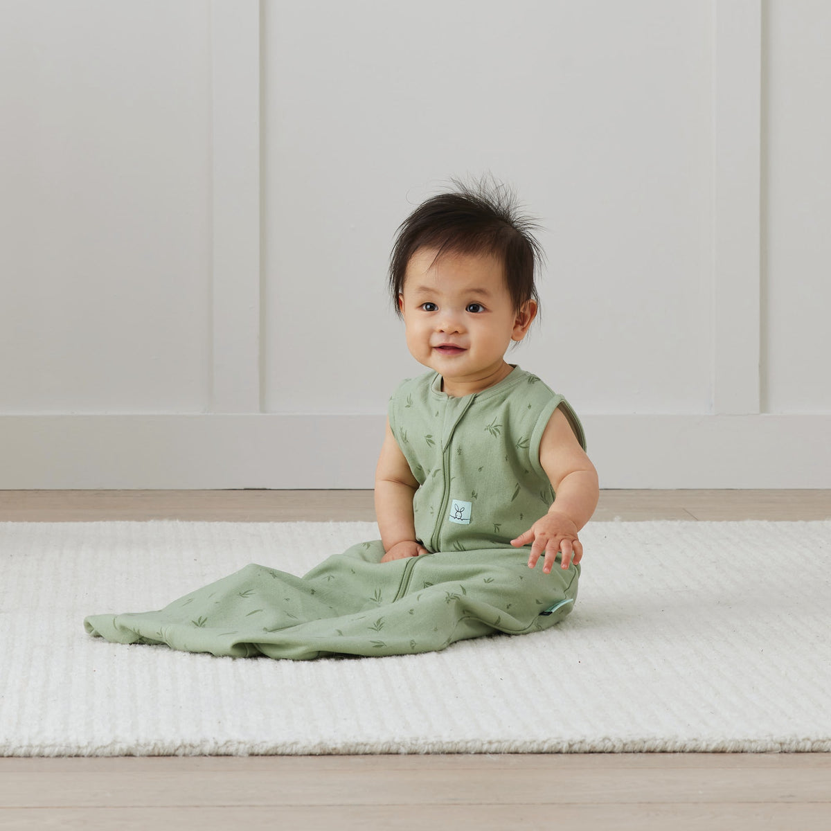 Baby in a green sleeping bag sitting on a white mat with a white wall background